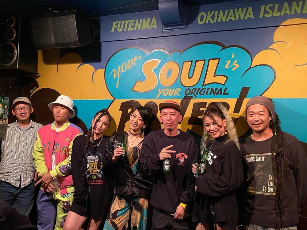 seven reggae lovers from Fukuoka and okinawa standing in line with whole-wall wall paint done by Massa AquaFlow as background for taking photo.